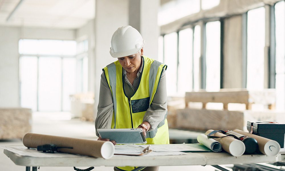 Blog - Woman Contractor Looking at Tablet in a Building at a Table with Documents